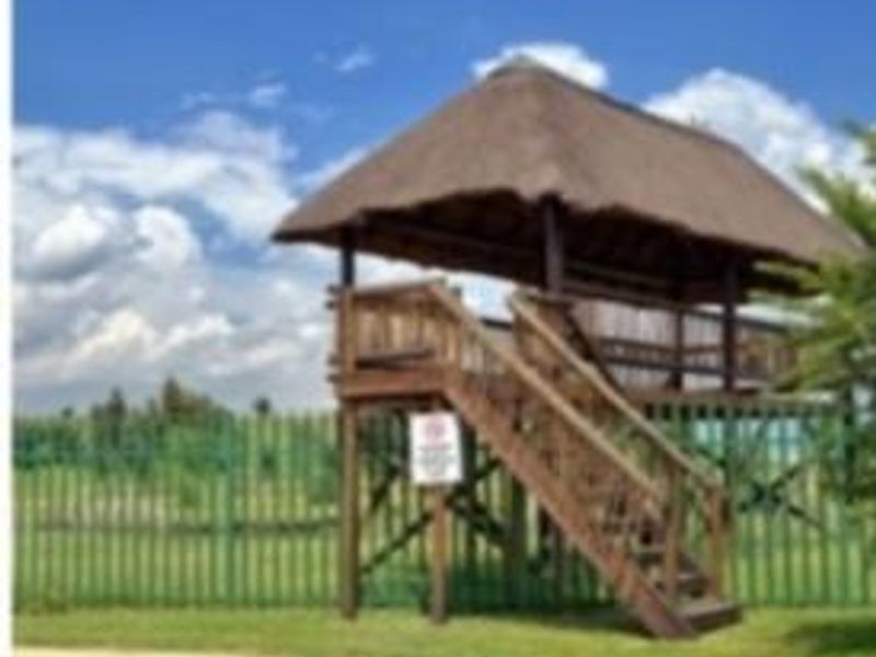A raised thatched gazebo structure with wooden stairs, situated within a landscaped outdoor area featuring perimeter fencing.