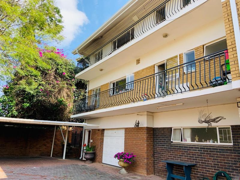Exterior view of a multi-story apartment building featuring brick facade, private balconies with iron railings, a garage, and a paved driveway with carport.