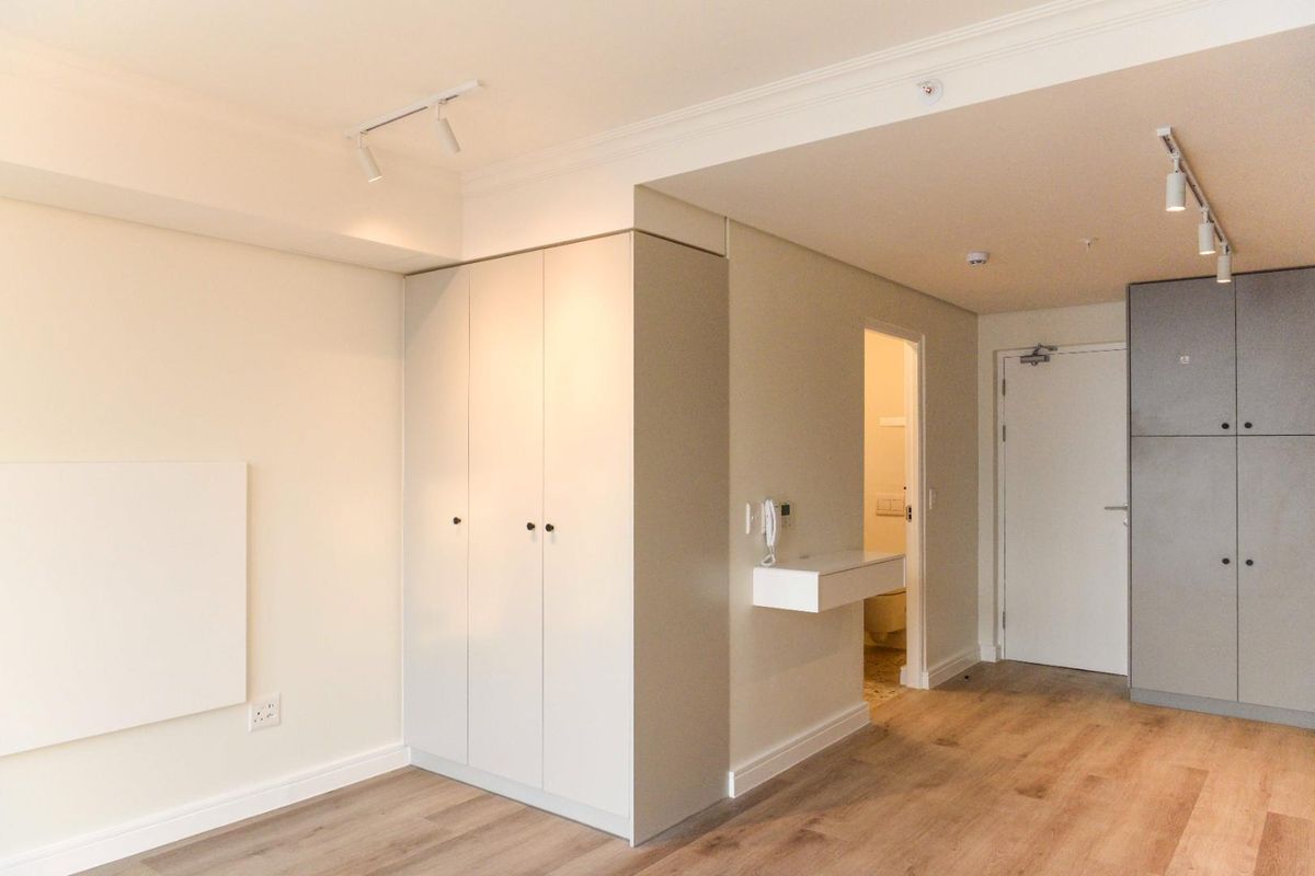 A bright, modern bedroom featuring neutral tones, laminate flooring, built-in storage, and track lighting, with an adjacent en-suite bathroom.