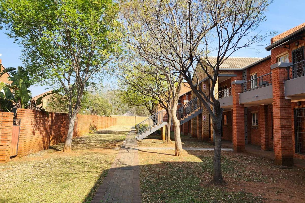 A view of the apartment complex rear exterior, featuring a paved walkway, mature trees, and facebrick building facades with external staircases.
