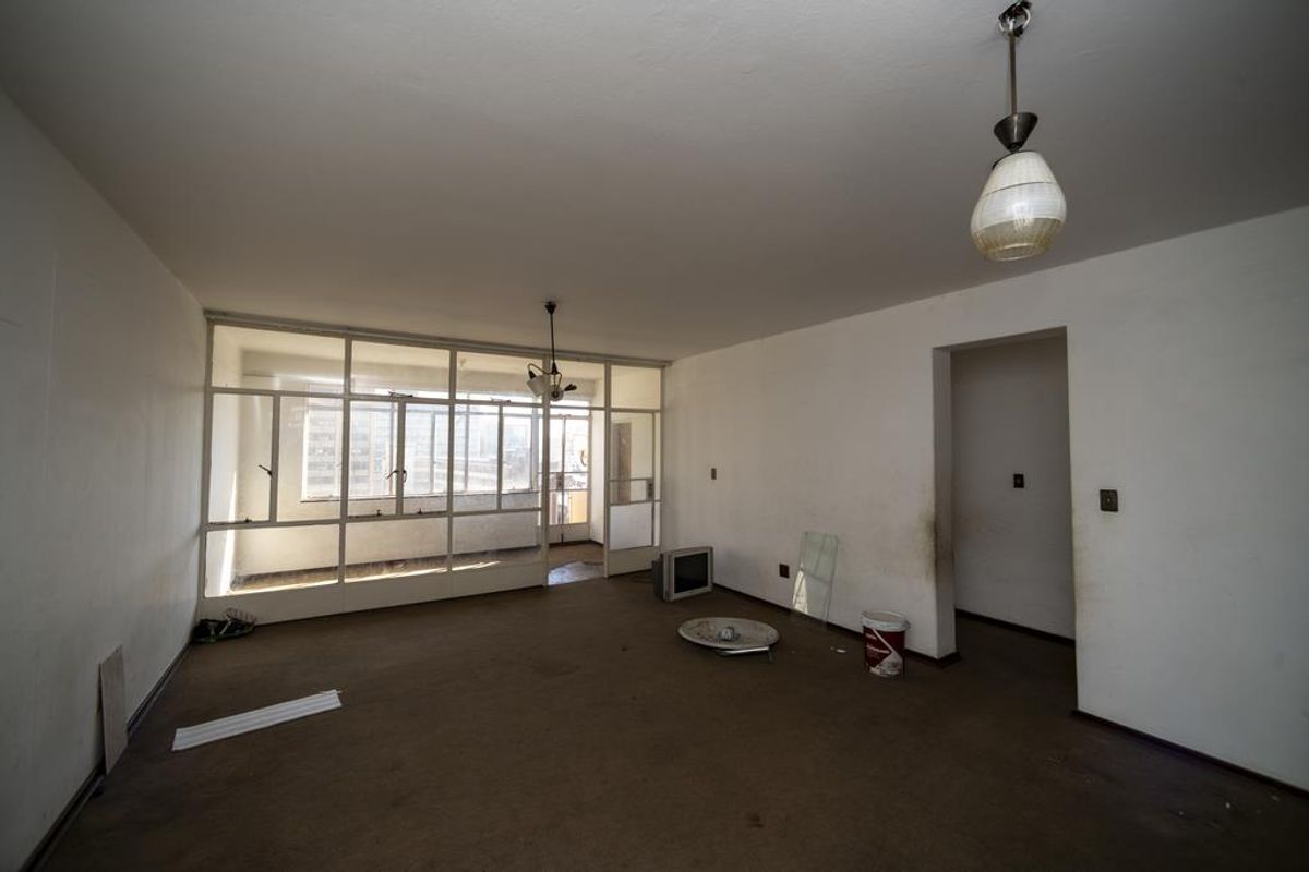 An empty, dated living area featuring a large glass-partitioned sunroom, original light fixtures, and worn carpeting throughout the space.