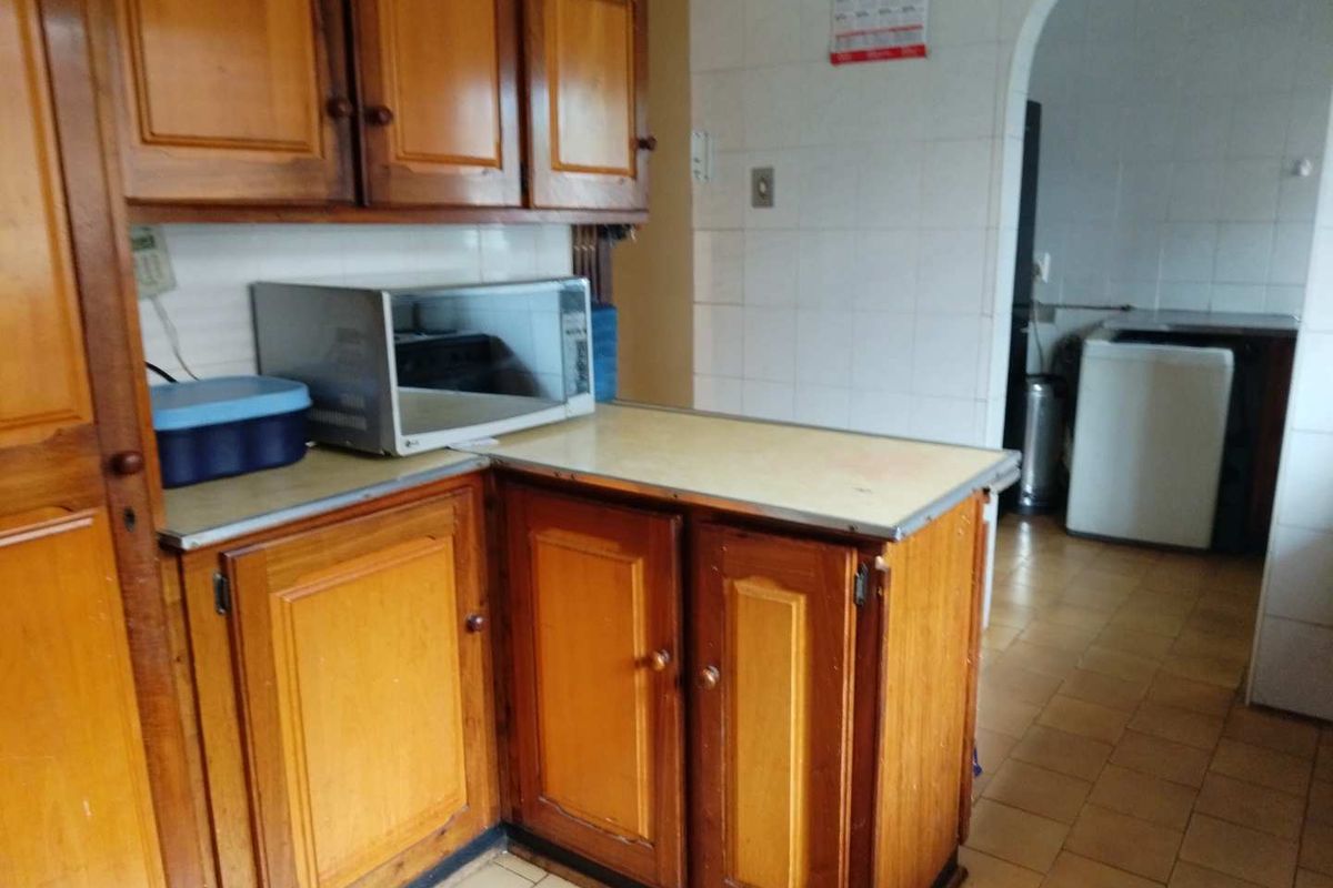A dated kitchen featuring traditional wooden cabinetry, tiled walls, and a tiled floor, with an adjacent scullery area visible through the arched doorway.
