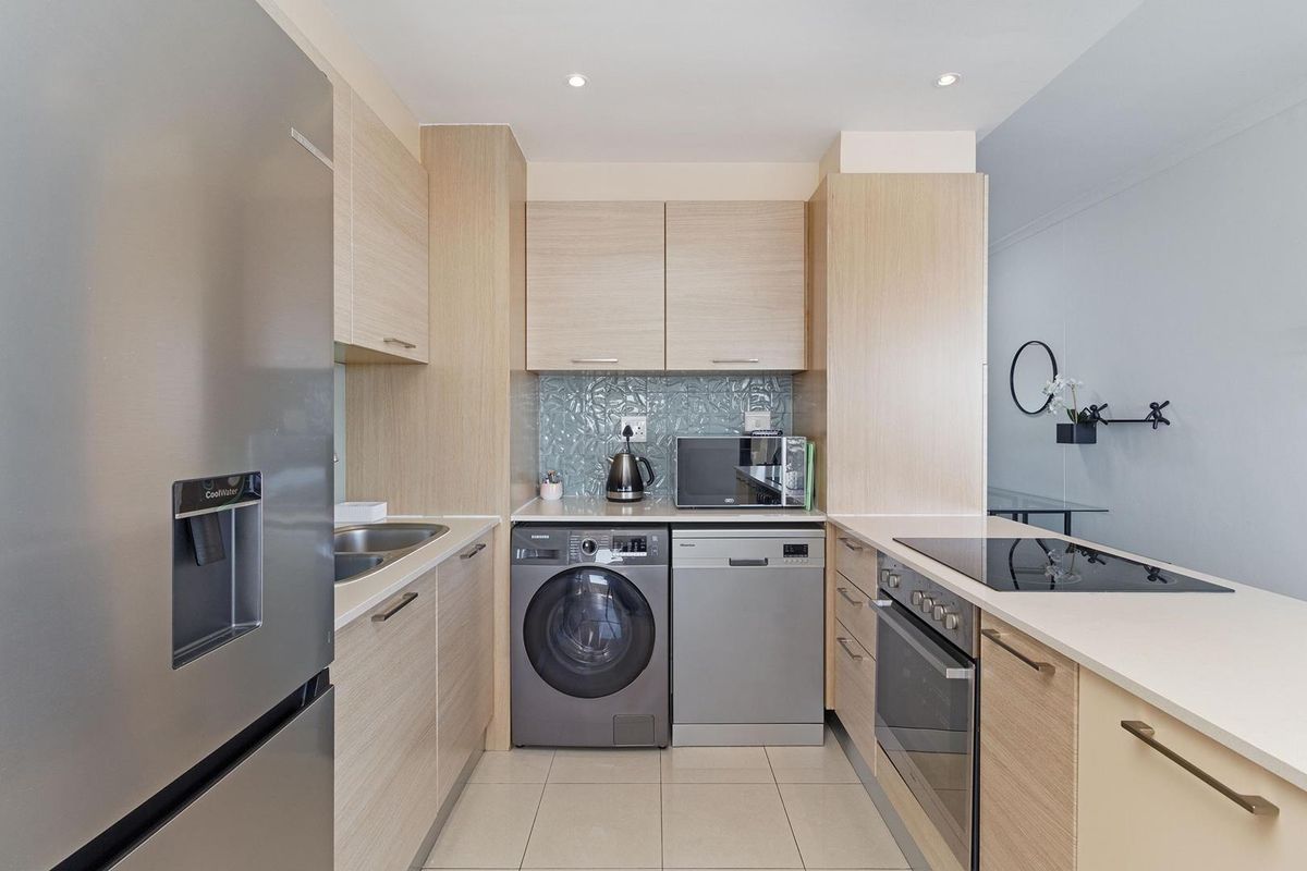 Modern galley-style kitchen featuring light wood cabinetry, stone countertops, integrated appliances, and a stylish textured glass backsplash.