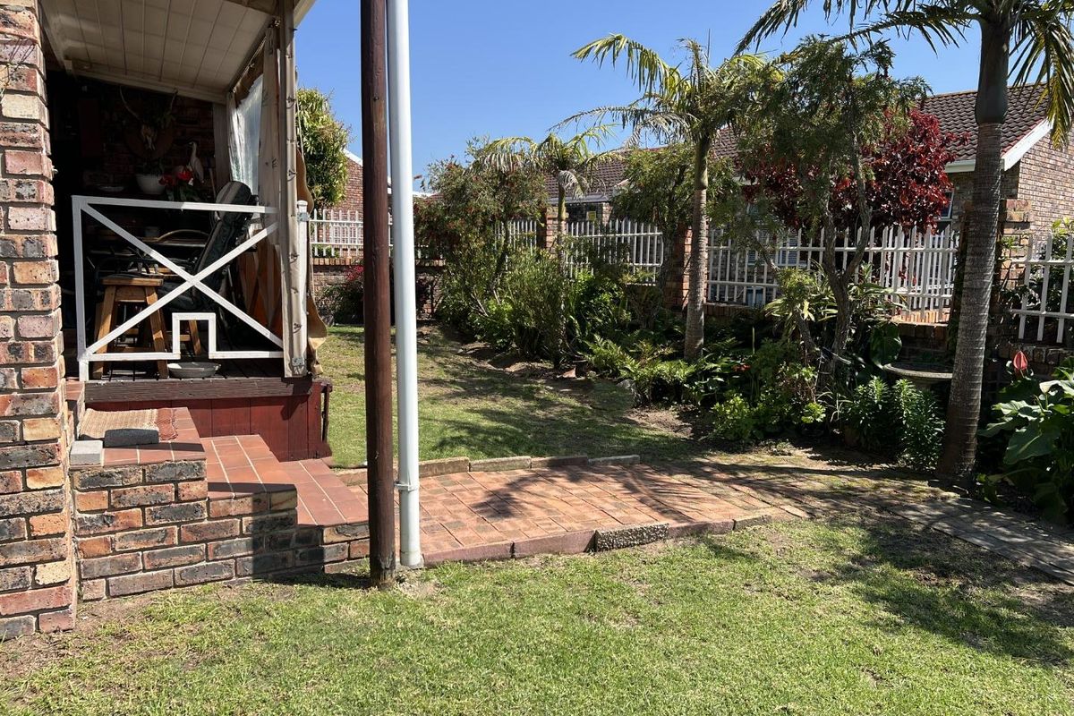 A rear exterior view of a townhouse featuring a covered patio, brick facade, paved walkway, and a landscaped garden with perimeter fencing.