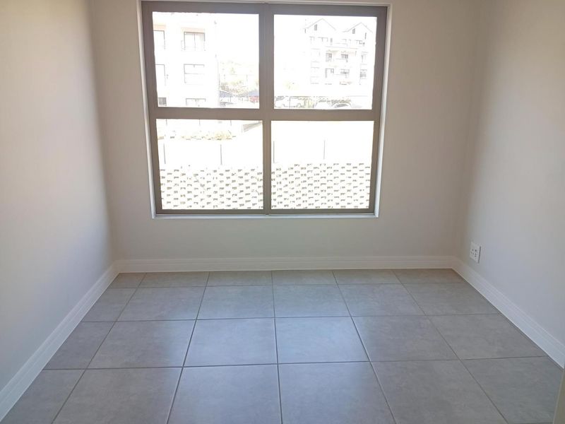 A bright, empty bedroom featuring neutral tiled flooring, clean white walls, and a large window providing natural light.