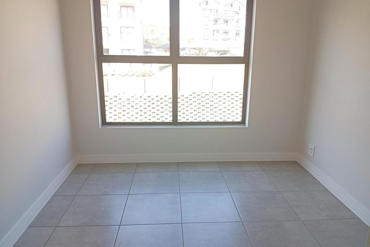 A bright, empty bedroom featuring neutral tiled flooring, clean white walls, and a large window providing natural light.