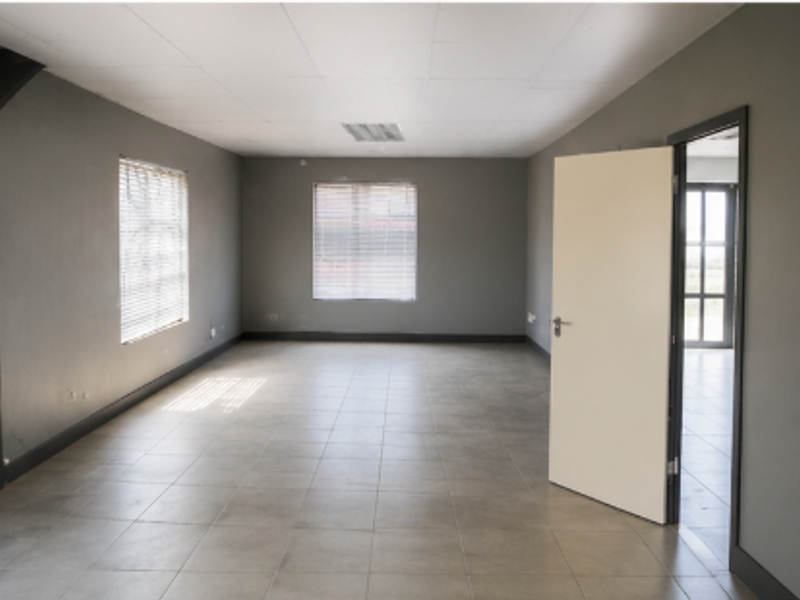 A spacious, empty office area featuring neutral grey walls, tiled flooring, and a drop ceiling with integrated lighting.