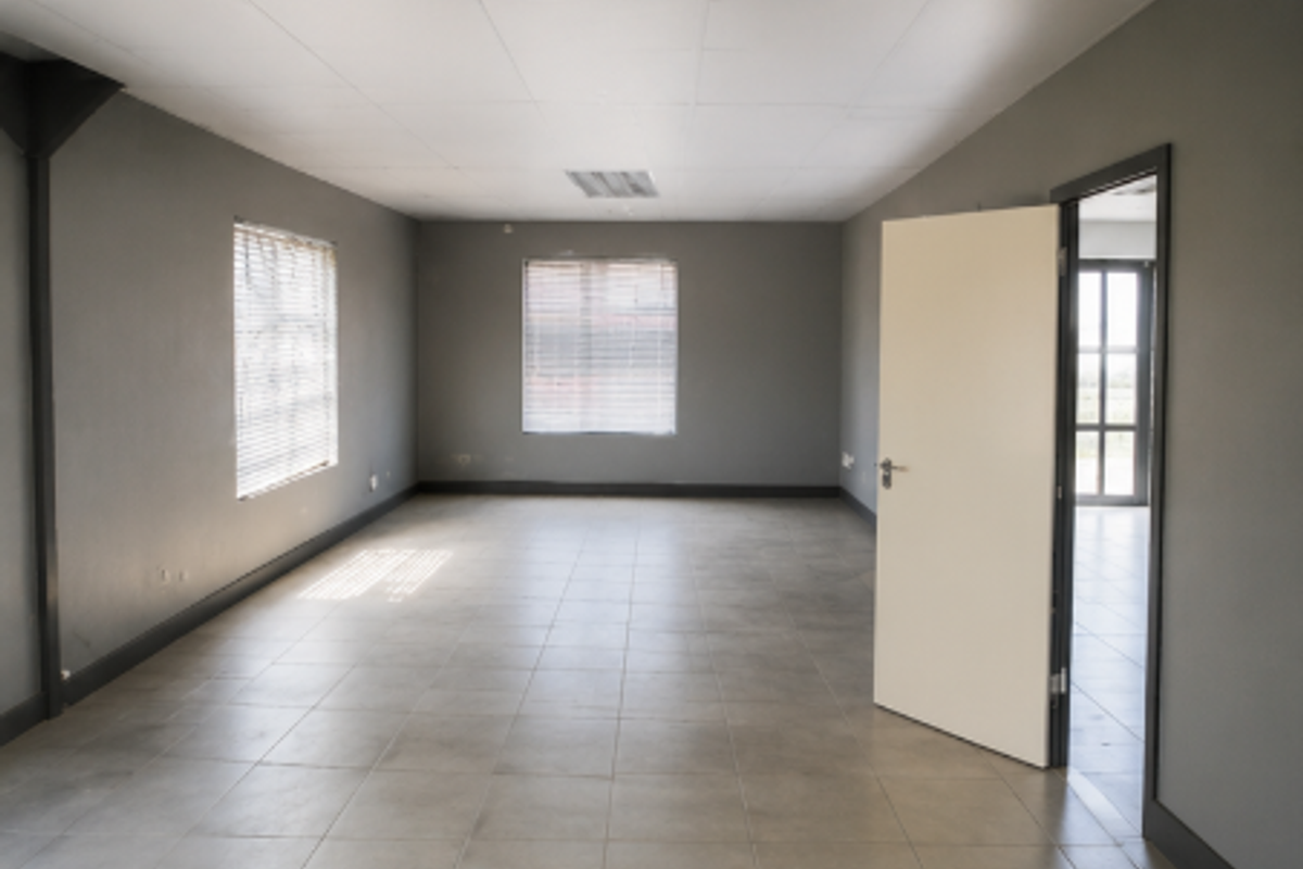 A spacious, empty office area featuring neutral grey walls, tiled flooring, and a drop ceiling with integrated lighting.