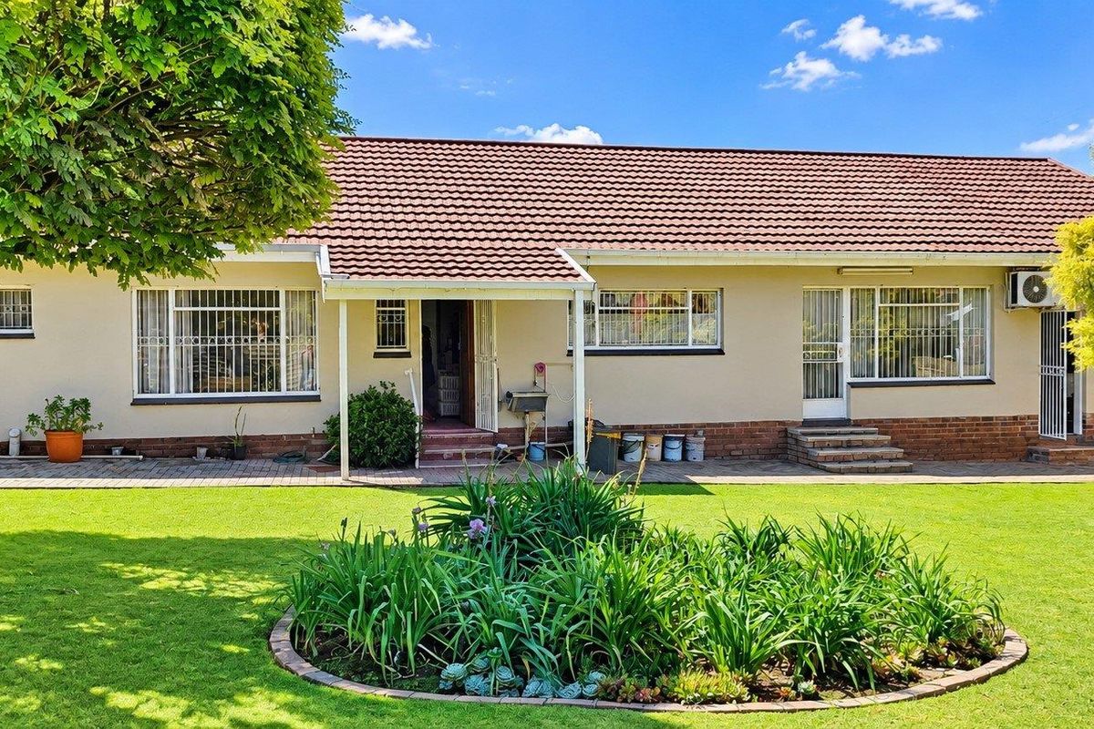 A well-maintained single-story house featuring a tiled roof, brick base, security bars on windows, and a spacious rear garden with a paved patio area.