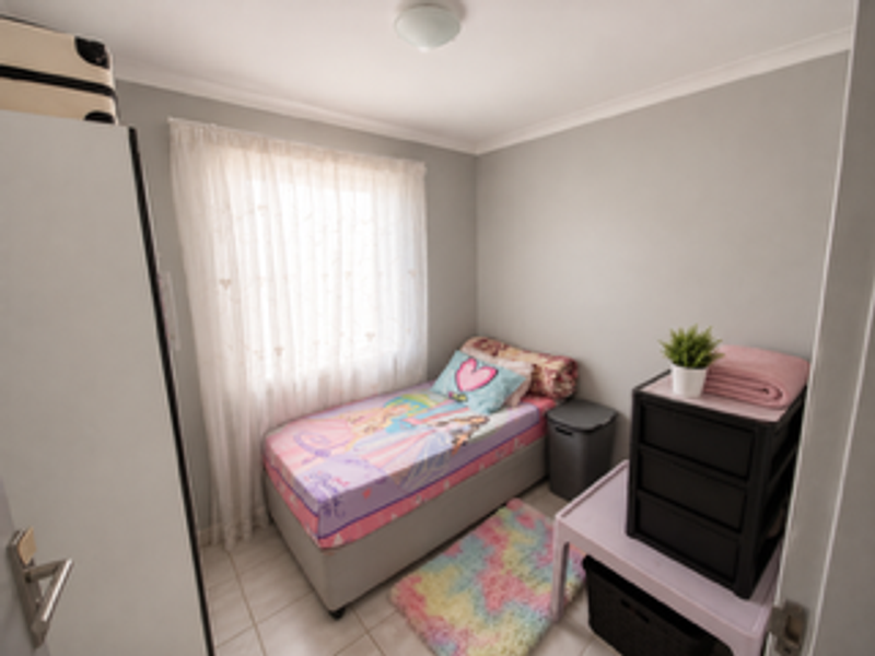 A compact bedroom featuring tiled flooring, a built-in cupboard, and a window with sheer curtains, providing a functional and well-lit space.
