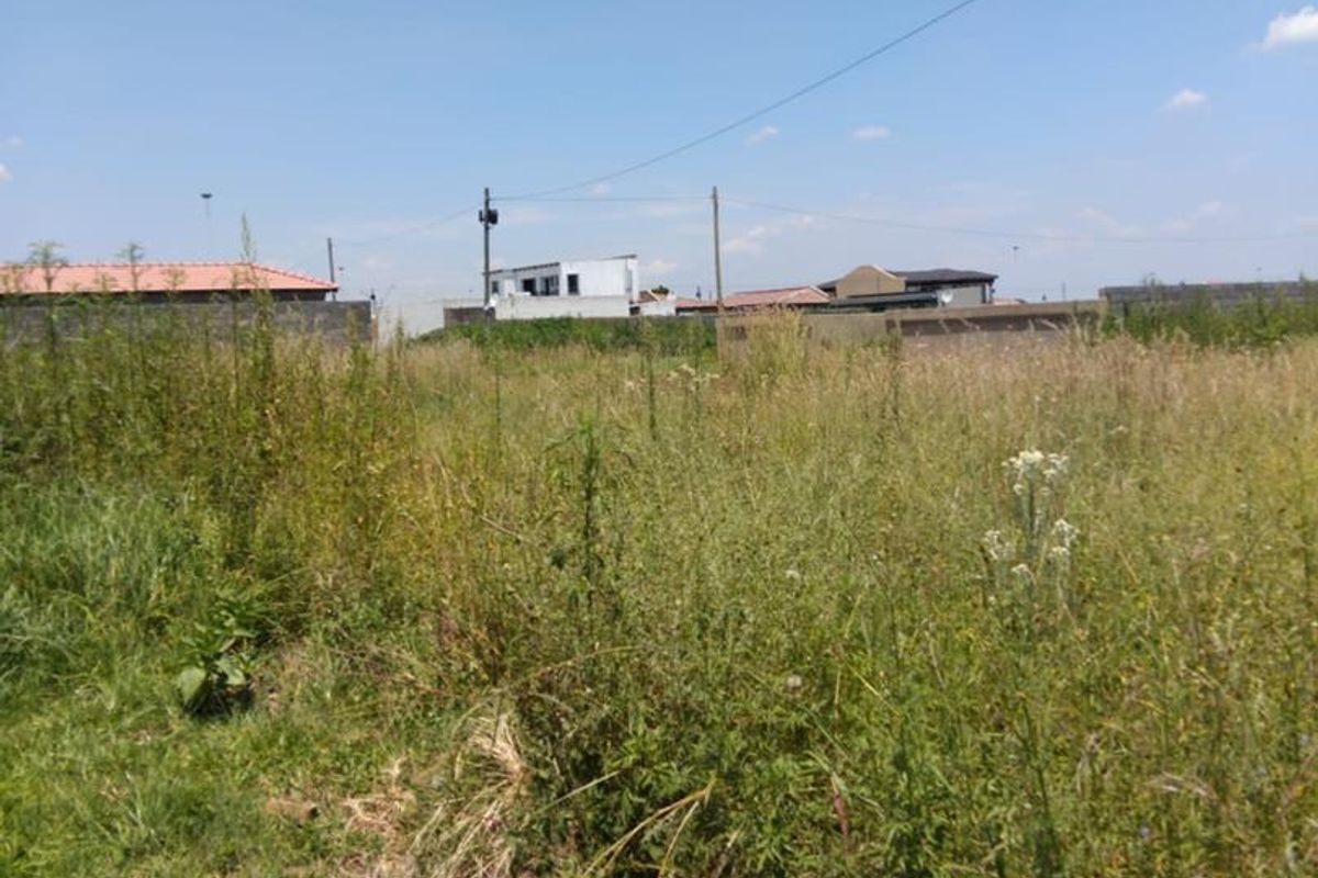 A view of a vacant residential plot covered in overgrown grass and vegetation, with neighboring properties visible in the background.