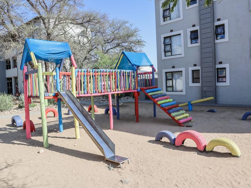 Communal outdoor playground area located at the rear of the apartment complex, featuring fixed play structures and sandy ground surface.