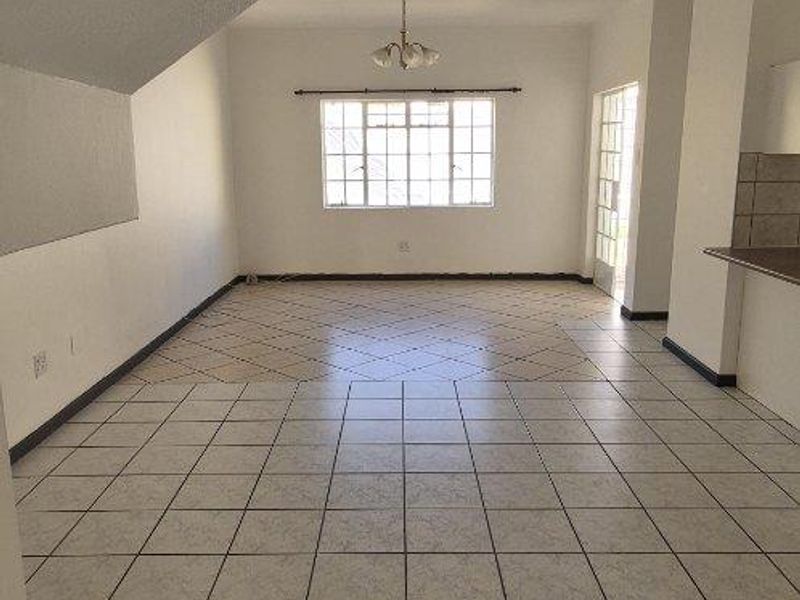 An empty open-plan living area featuring ceramic floor tiles, a staircase underside, and natural light from a rear window and door.