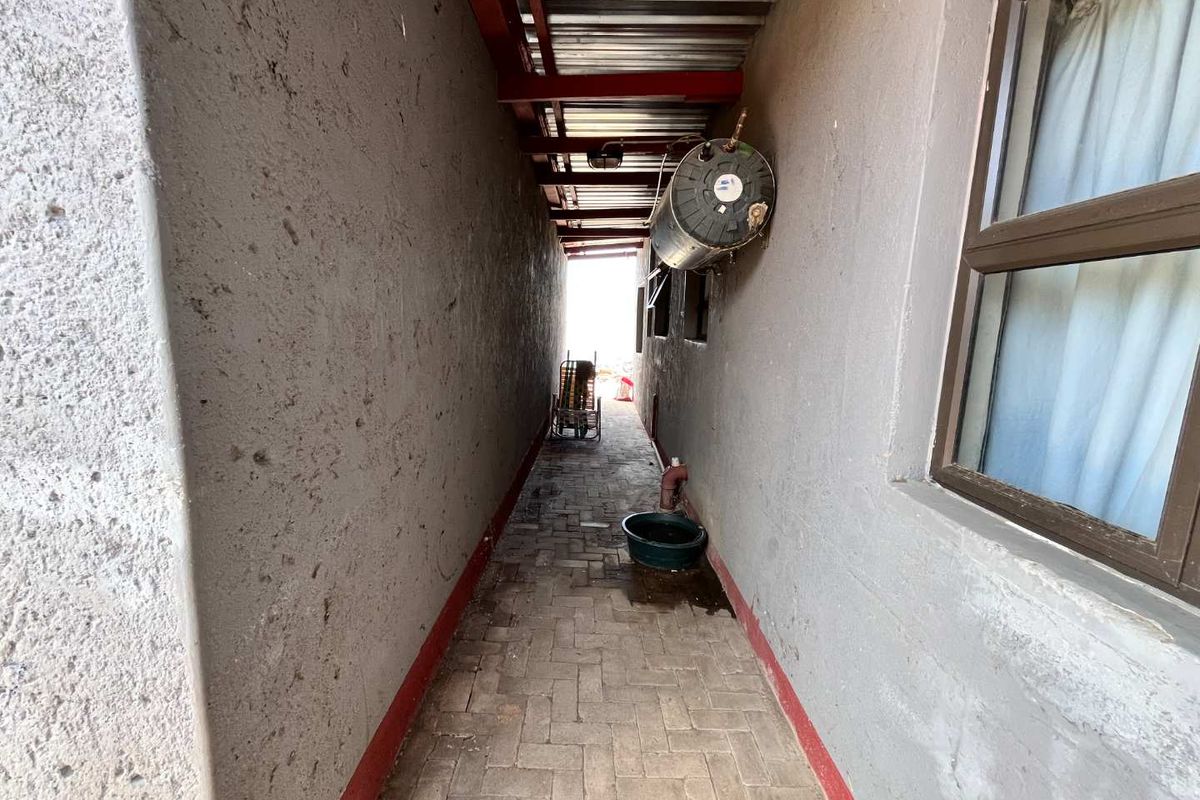 A narrow exterior side passage featuring paved flooring, a wall-mounted electric geyser, and a corrugated metal roof covering.