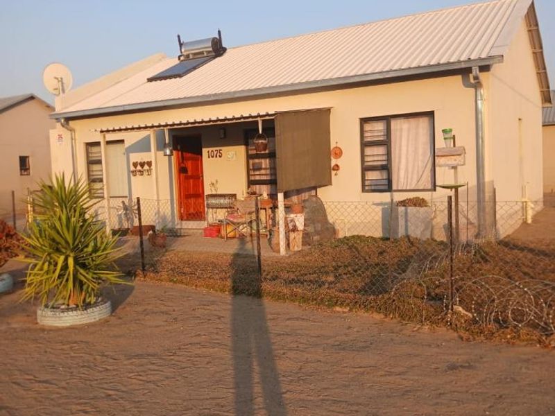 Front view of a single-story house featuring a corrugated metal roof, solar water heater, and a fenced yard with a covered porch entrance.