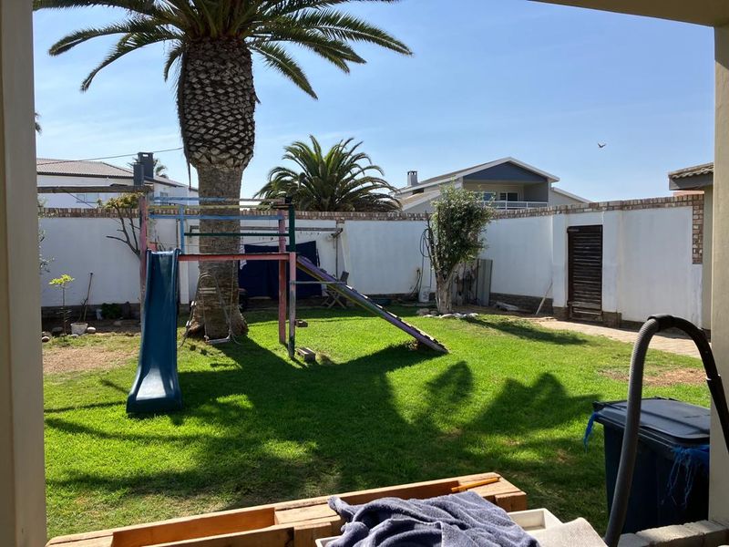 A private rear garden featuring a lawn, mature palm tree, and paved walkway, enclosed by a boundary wall and viewed from a covered patio.