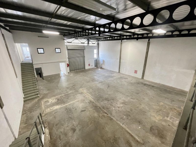 Spacious industrial warehouse unit featuring high ceilings with exposed steel trusses, concrete flooring, and a large roller shutter door for easy access.