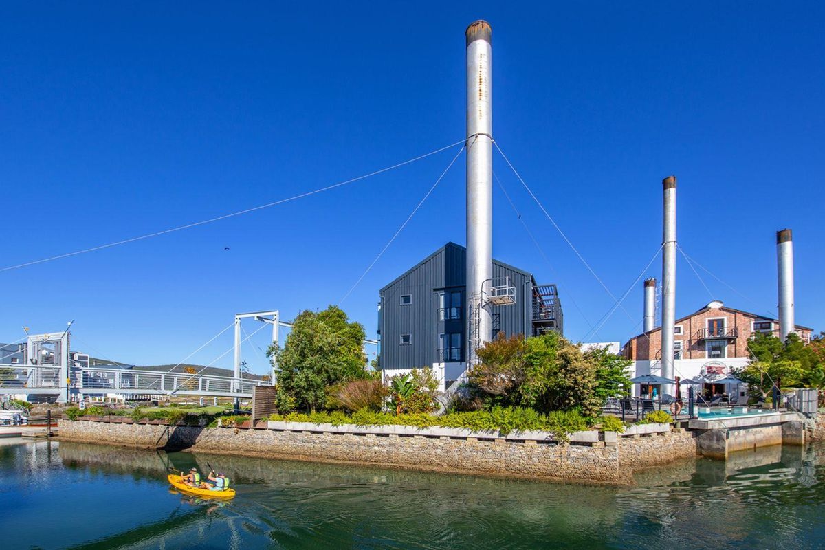 A scenic waterfront view of a unique industrial-style building complex featuring tall chimneys, landscaped grounds, and a swimming pool area.