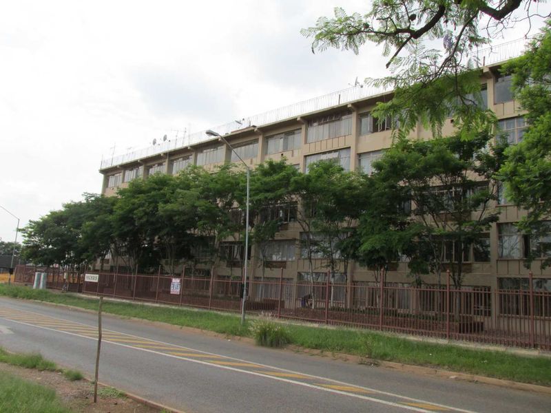 Exterior view of a multi-story apartment building featuring a metal perimeter fence and mature trees along the street frontage.
