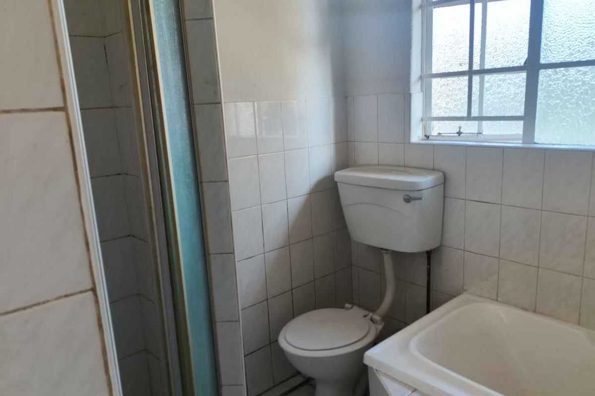A dated bathroom featuring a shower enclosure, bathtub, and toilet with tiled walls and a window with security bars.