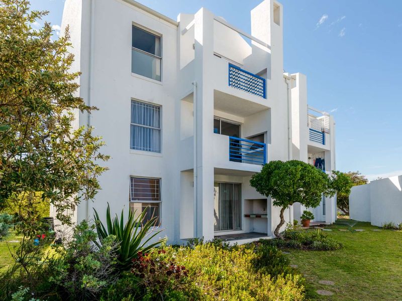 Rear view of a multi-story apartment building featuring private balconies, sliding glass doors, and a built-in braai area overlooking a landscaped garden.