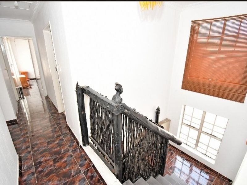 A bright hallway featuring tiled flooring, a unique wooden balustrade, and natural light through grid windows with venetian blinds.