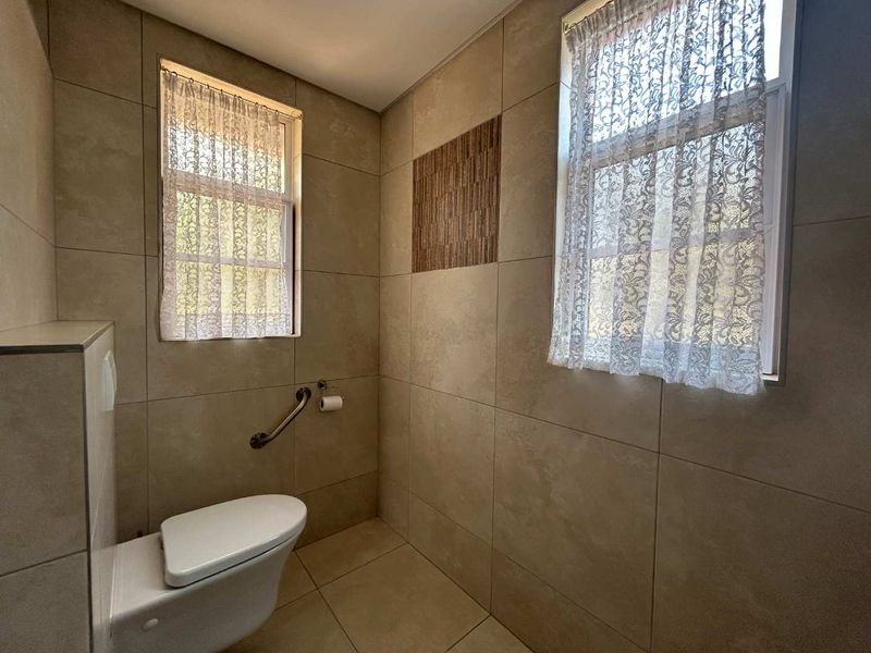 A clean, neutral-toned bathroom featuring floor-to-ceiling tiling, a wall-mounted toilet, and a safety grab rail, offering a functional and accessible space.