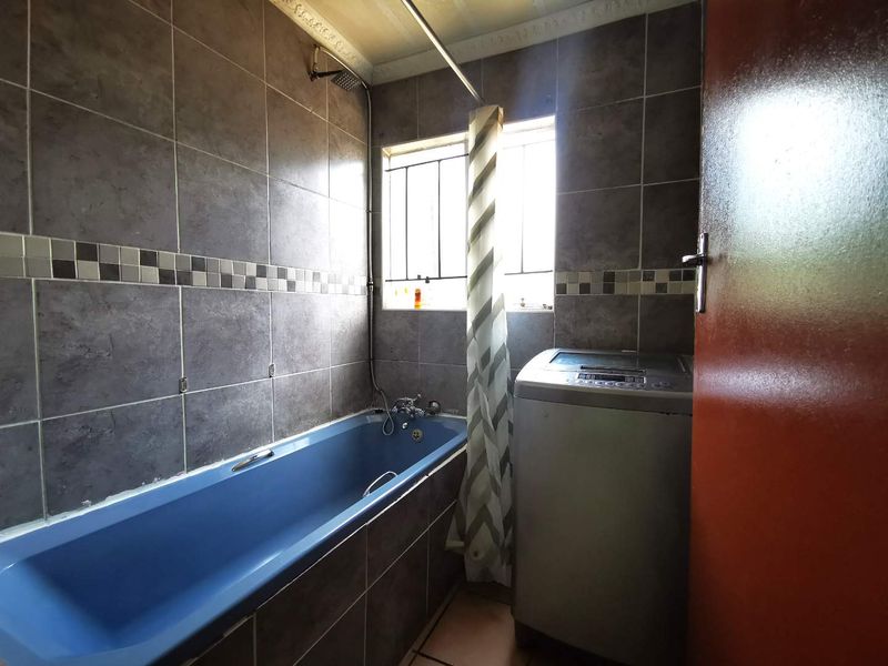 A functional bathroom featuring dark grey wall tiles, a blue bathtub with integrated shower, and security bars on the window.