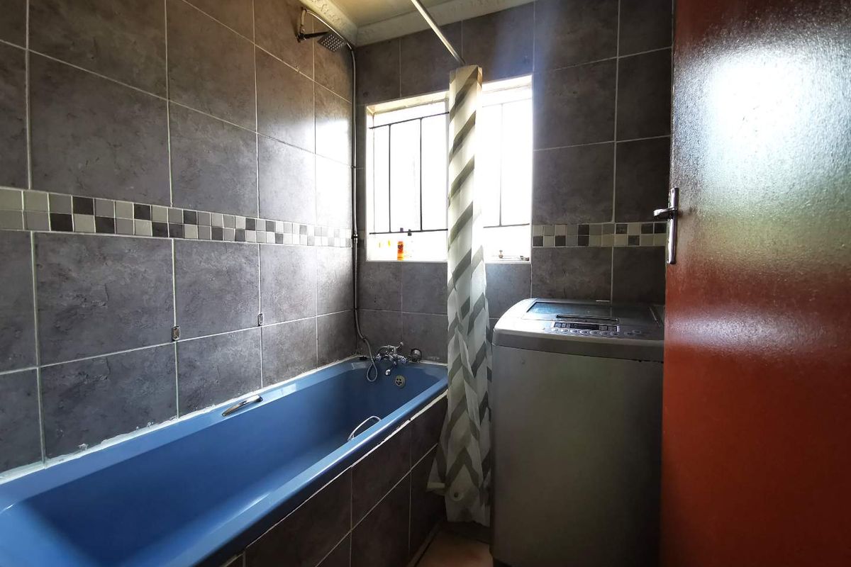 A functional bathroom featuring dark grey wall tiles, a blue bathtub with integrated shower, and security bars on the window.