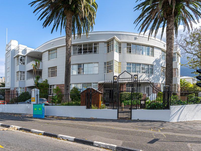 Street-level view of a classic curved apartment building featuring distinctive architectural styling, wrought iron fencing, and mature palm trees.