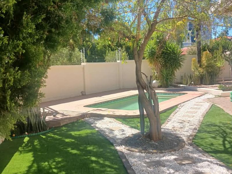 A well-maintained outdoor pool area featuring a swimming pool, paved surround, landscaped garden, and secure perimeter wall with electric fencing.