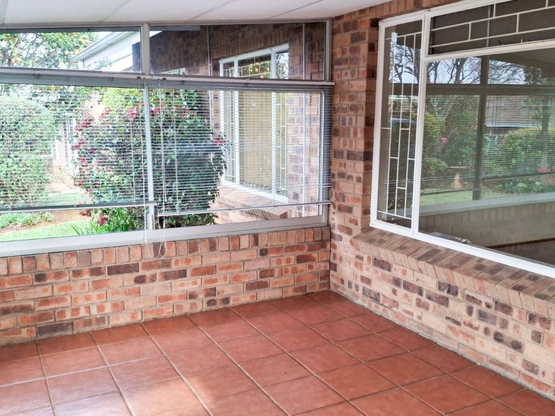 A sun-filled enclosed patio featuring face-brick walls, tiled flooring, and large aluminium-framed windows overlooking the garden.