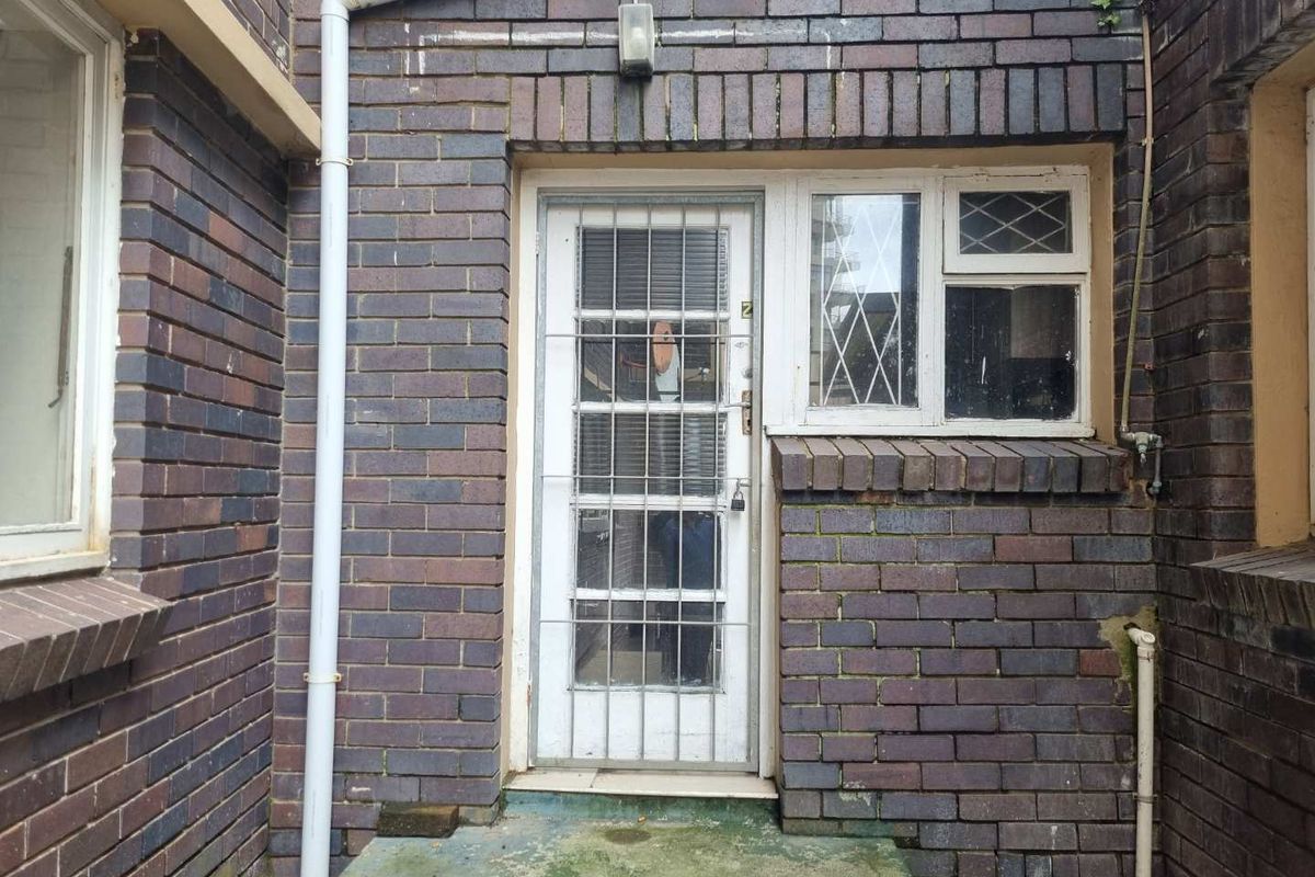 Rear exterior view of the apartment featuring dark brickwork, a security gate, and a concrete step entrance.