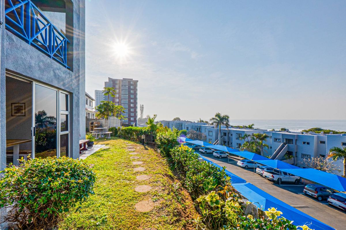An elevated view of the apartment complex exterior, featuring a private garden area, balcony access, and shaded parking facilities with ocean views.
