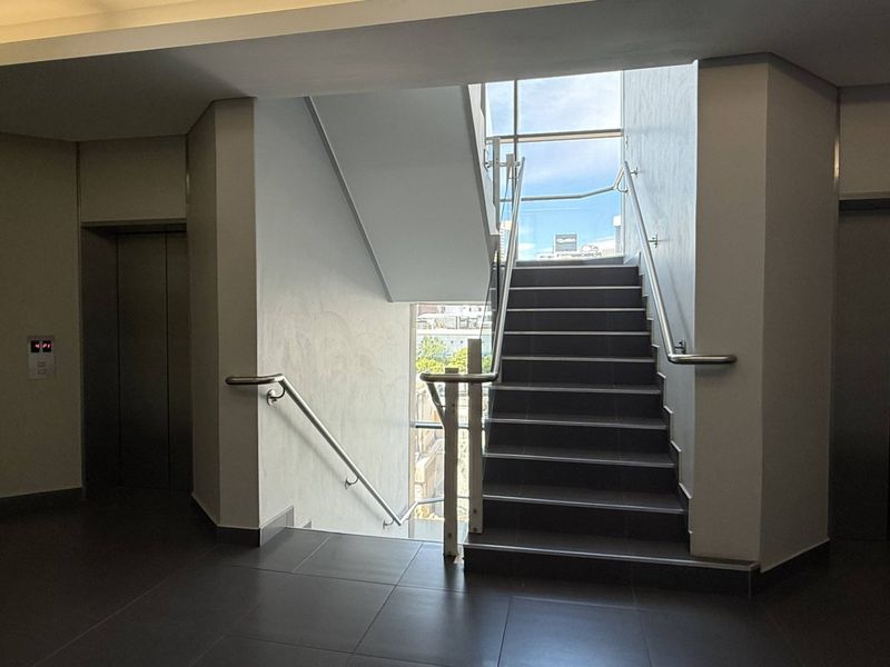 Modern office building lobby featuring a central staircase with glass balustrades, stainless steel handrails, and dual elevator access.