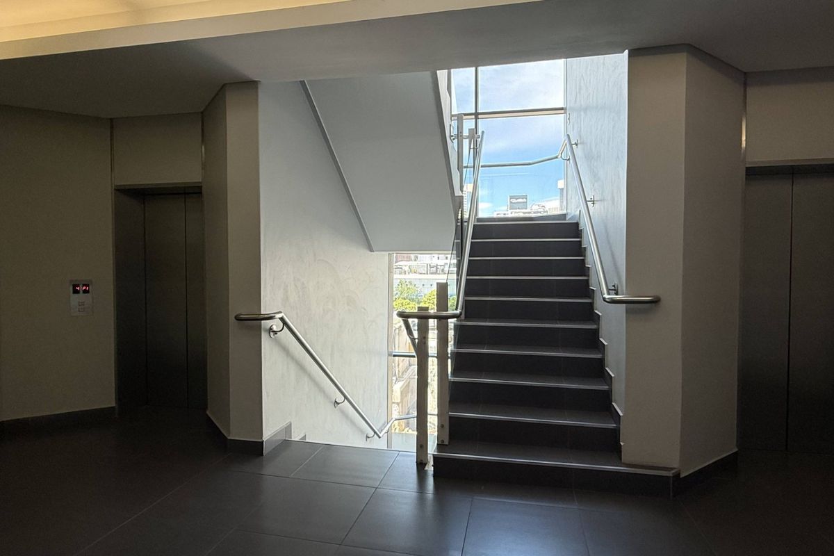 Modern office building lobby featuring a central staircase with glass balustrades, stainless steel handrails, and dual elevator access.