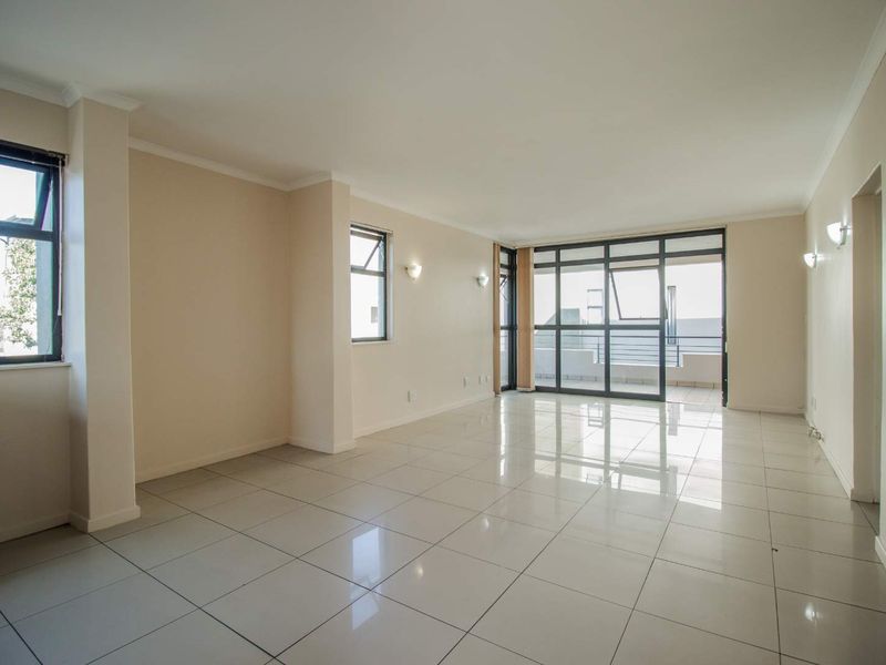 A spacious, bright, and empty living room featuring neutral tiled flooring, modern aluminium window frames, and a sliding glass door leading to a balcony.