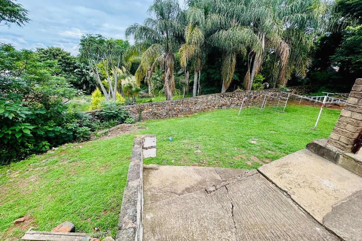 A rear garden area featuring a lawn, mature trees, a stone boundary wall, and a concrete patio with visible structural cracking.