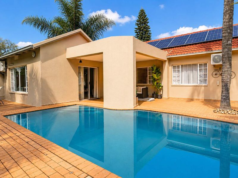 A well-maintained outdoor area featuring a swimming pool, brick-paved surround, wooden decking, and a covered patio, complemented by roof-mounted solar panels.