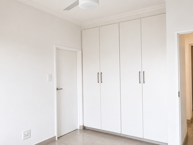 A clean, bright bedroom featuring neutral tiled flooring, white built-in cupboards, and a ceiling fan, offering a blank canvas for personalization.