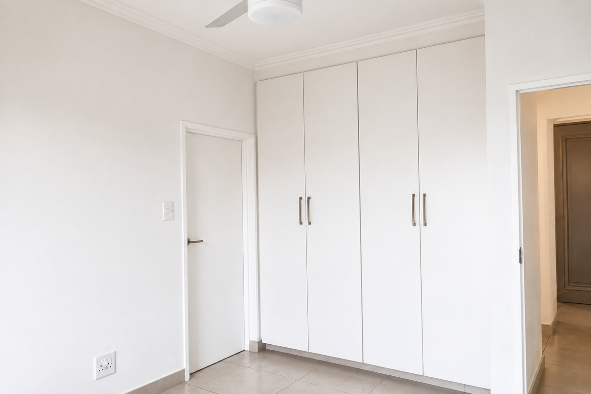 A clean, bright bedroom featuring neutral tiled flooring, white built-in cupboards, and a ceiling fan, offering a blank canvas for personalization.