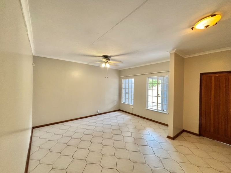 A spacious, empty living area featuring neutral walls, tiled flooring, a ceiling fan, and natural light through grid-style windows.