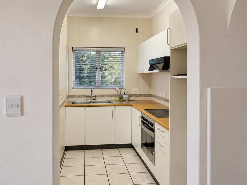 A functional kitchen featuring white cabinetry, laminate countertops, built-in oven and hob, tiled splashbacks, and tiled flooring, viewed through an arched doorway.