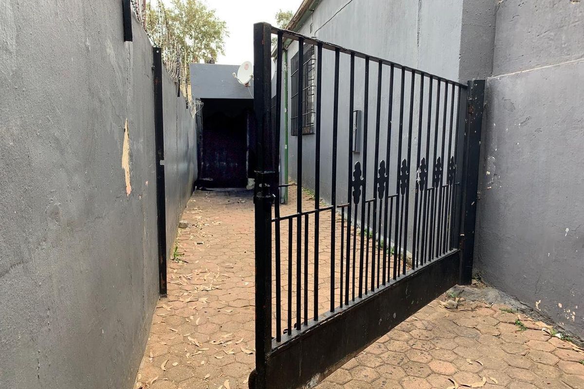 A narrow side passage featuring paved flooring, a metal security gate, and high boundary walls with security wire.