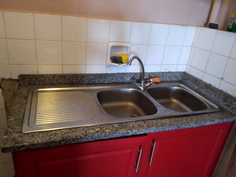 A kitchen sink area featuring a stainless steel double bowl sink, granite countertop, and tiled backsplash with visible wall damage on the left side.