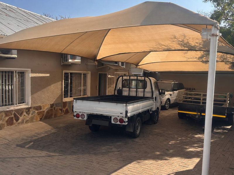 Rear exterior area featuring a large shade sail covering a paved parking space, stone cladding accents, and wall-mounted air conditioning units.