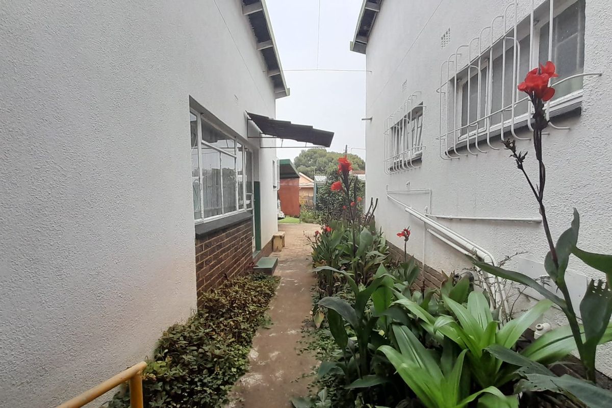 A narrow side walkway between two building sections, featuring a concrete path, exterior security bars on windows, and established garden beds.