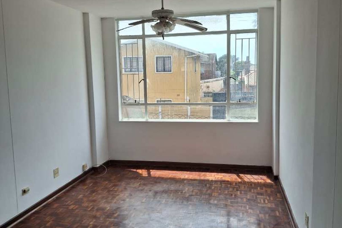 A bright, empty bedroom featuring classic parquet flooring, a ceiling fan, and large windows providing natural light and neighborhood views.