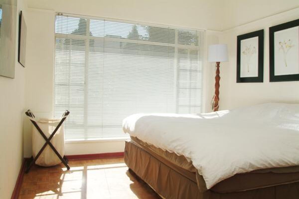 A bright bedroom featuring parquet flooring and large windows fitted with venetian blinds, offering a clean and functional living space.