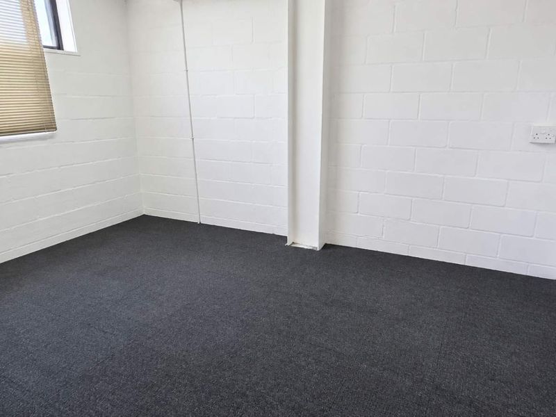 A clean, empty office space featuring painted concrete block walls, dark carpeting, and natural light from a window with fitted blinds.